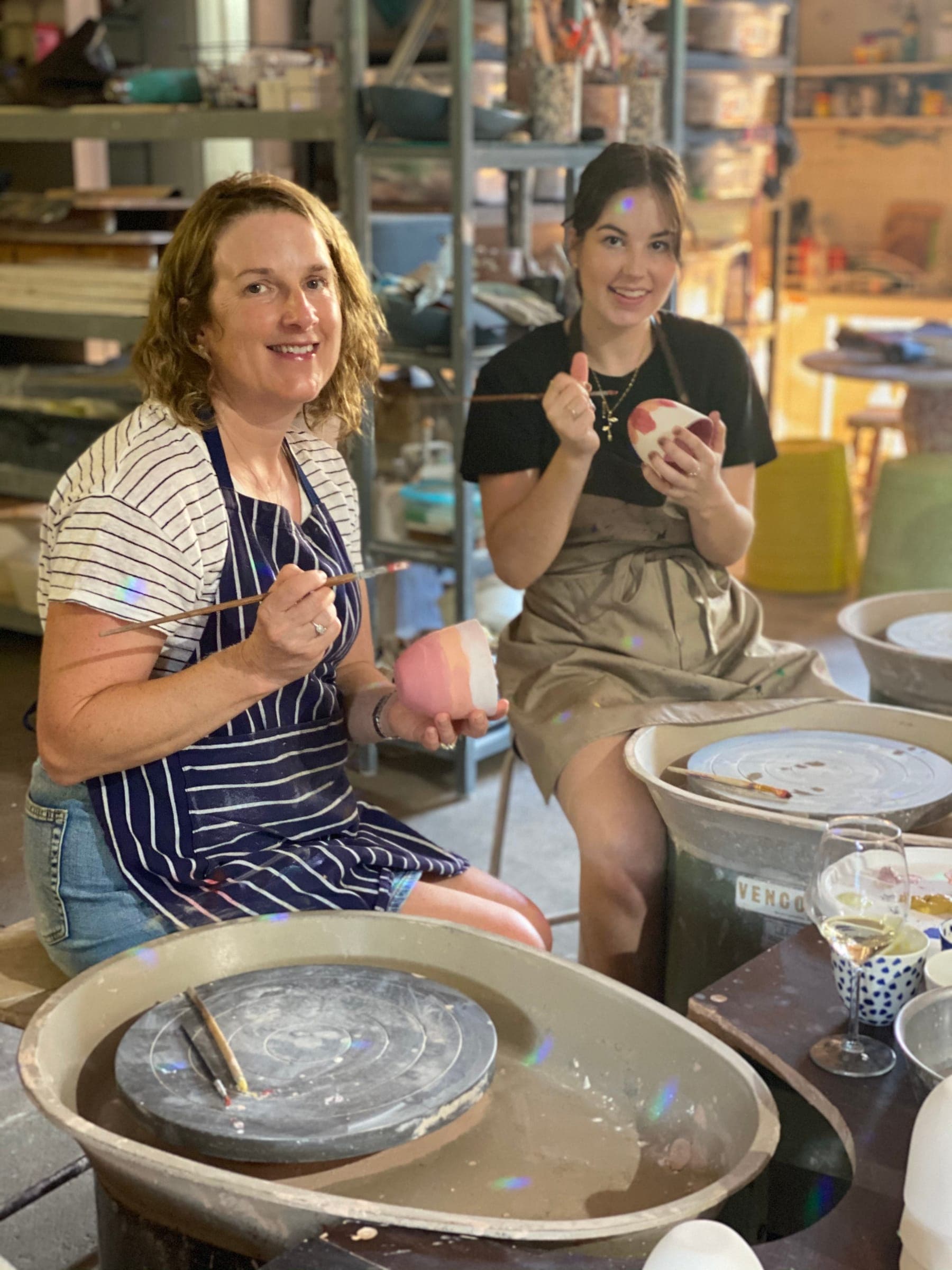 Two friends at the wheels, one holding up a freshly painted pink bowl