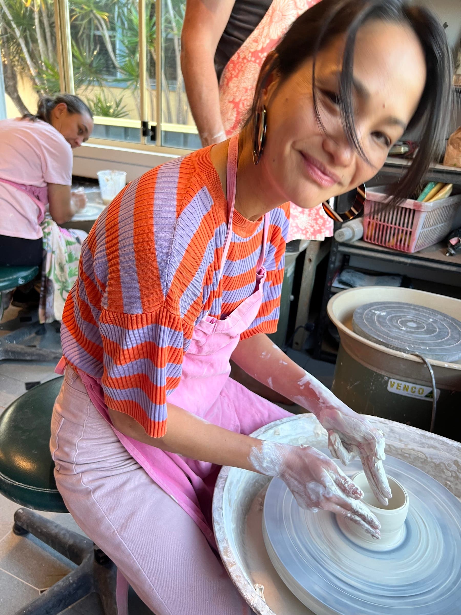 A maker in a striped jumper laughing with clay-covered hands at the wheel