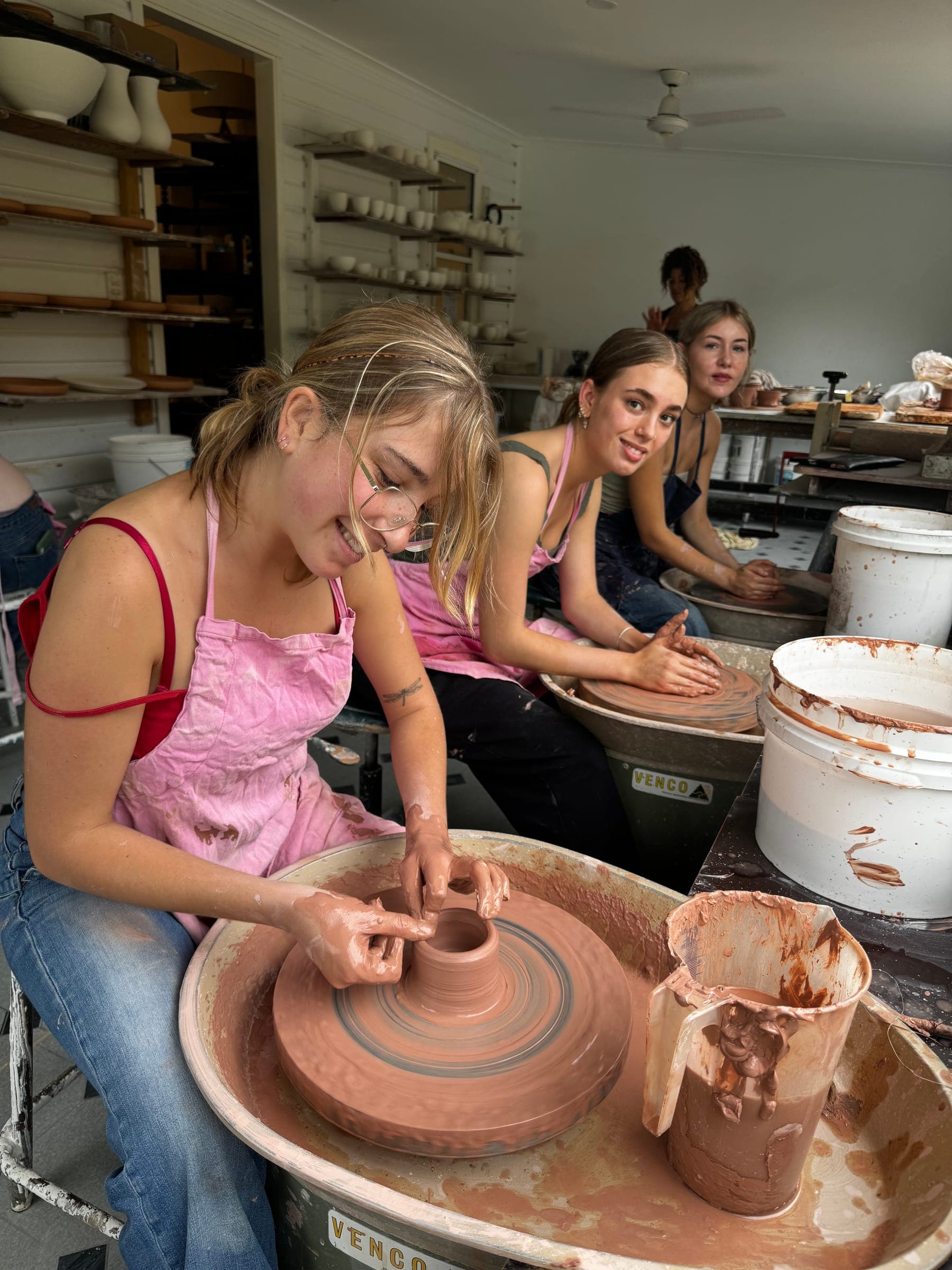 A row of students in pink aprons centring clay at the wheels